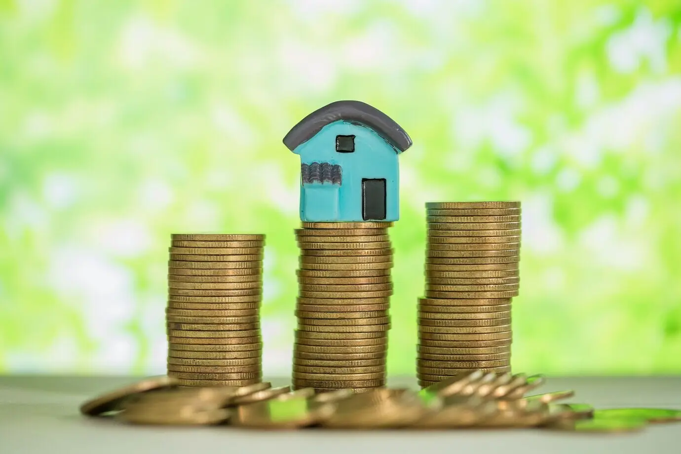 A miniature house atop a stack of coins, with a green blur.